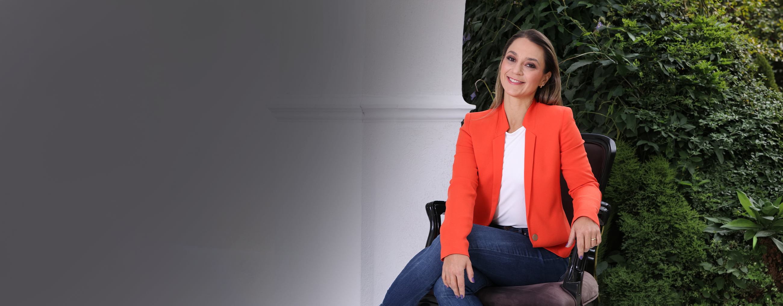 Ju Romantini sorrindo em frente a parede de plantas verdes, de blazer vermelho.